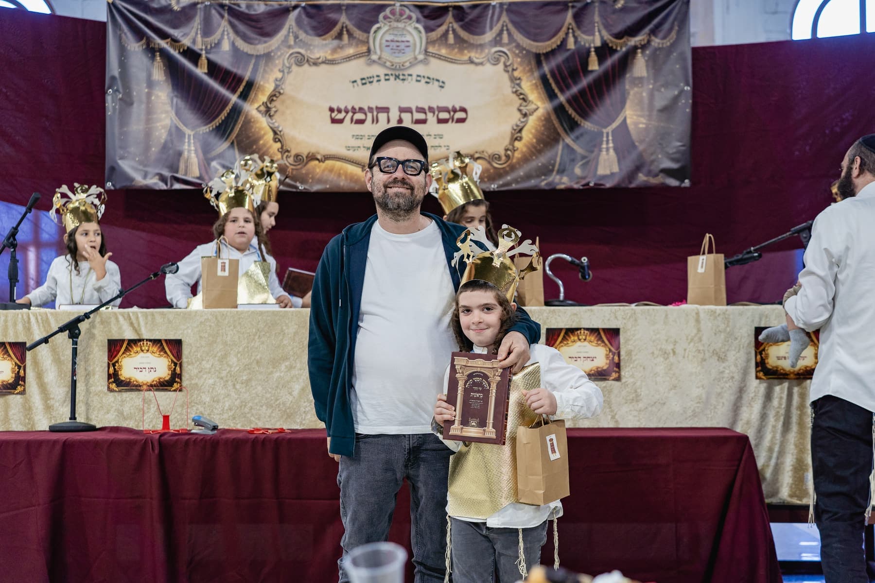 A student in a gold crown holding his Chumash beside a parent at Mesibat Chumash